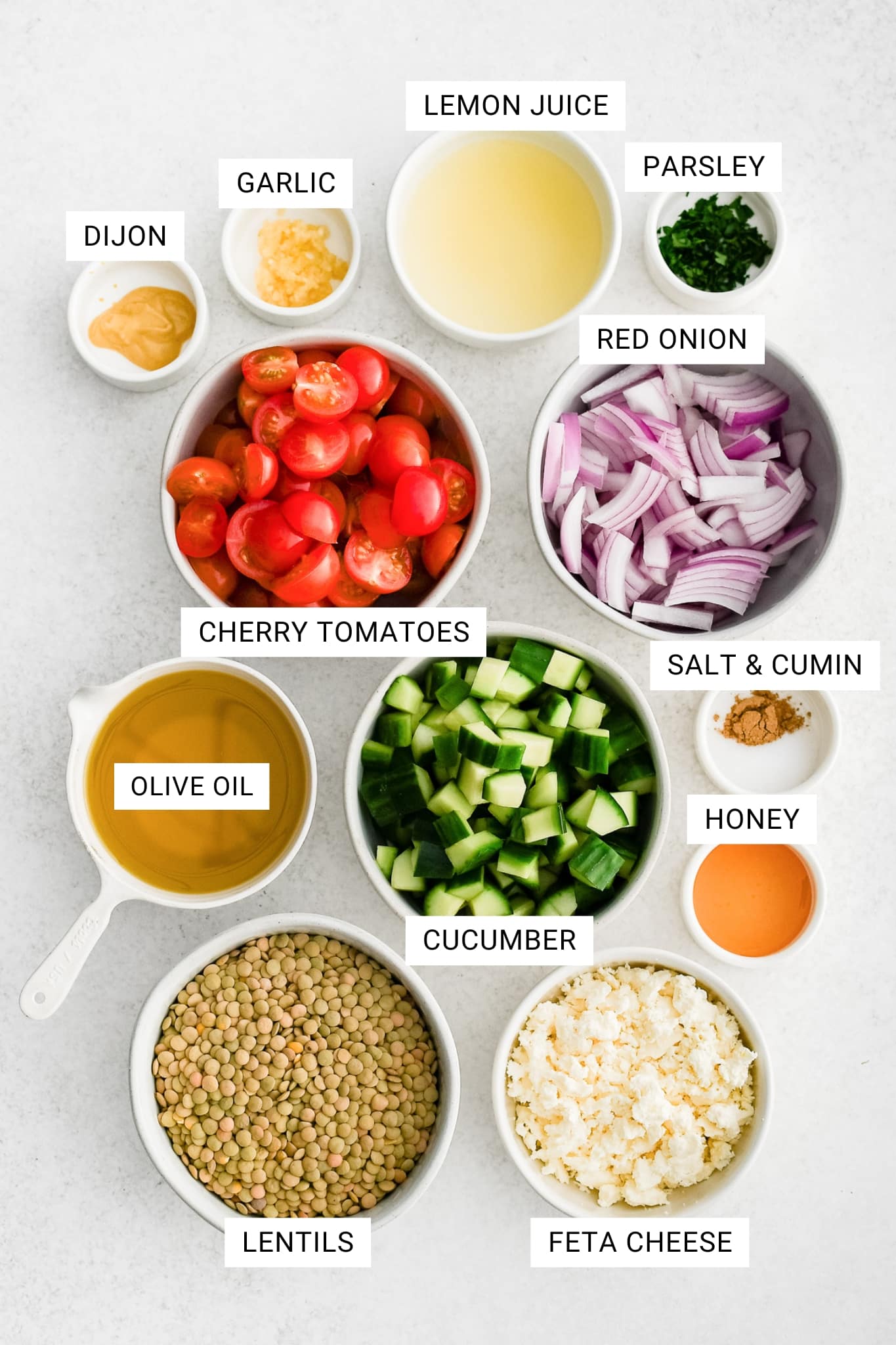 Overhead ingredients photo for lentil salad recipe arranged in white bowls and measuring cups on a light white background with text overlays labeling each ingredient: DIJON, GARLIC, LEMON JUICE, PARSLEY, RED ONION, CHERRY TOMATOES, OLIVE OIL, CUCUMBER, SALT & CUMIN, HONEY, LENTILS, and FETA CHEESE.