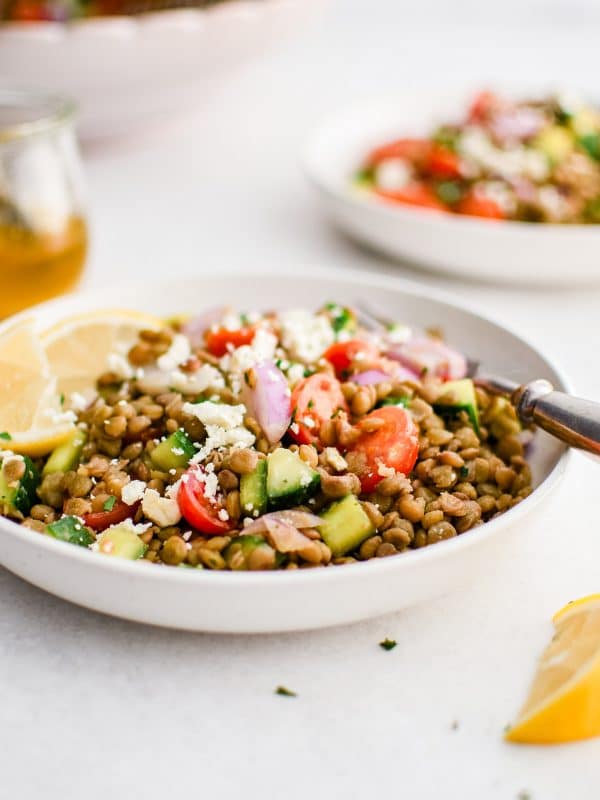 Side-angle photo of lentil salad served in a shallow white bowl with brown lentils, diced cucumber, halved cherry tomatoes, sliced red onion, crumbled feta cheese, and lemon wedges. A small glass jar of golden dressing sits to the left, with another bowl of salad blurred in the background.