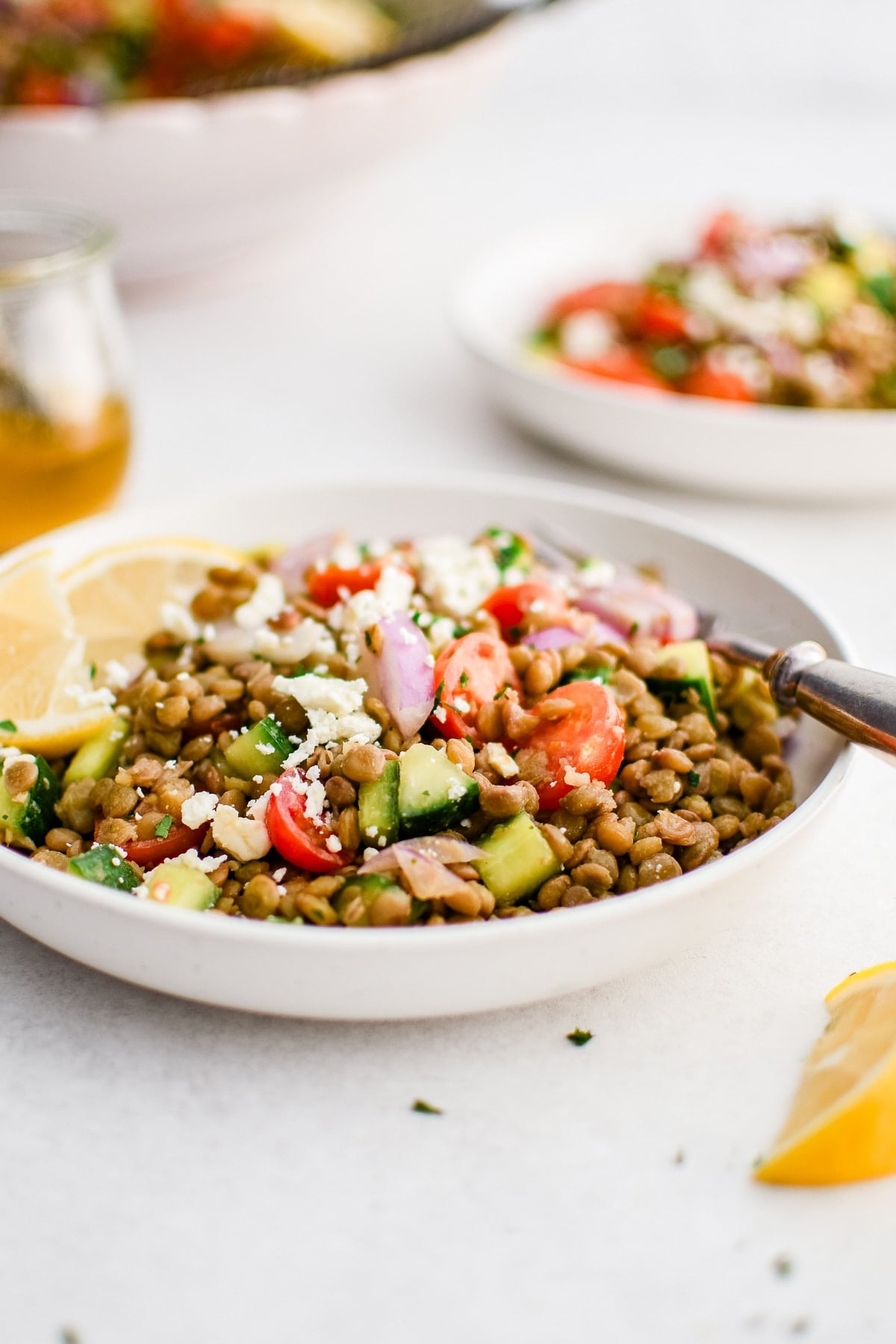 Side-angle photo of lentil salad served in a shallow white bowl with brown lentils, diced cucumber, halved cherry tomatoes, sliced red onion, crumbled feta cheese, and lemon wedges. A small glass jar of golden dressing sits to the left, with another bowl of salad blurred in the background.