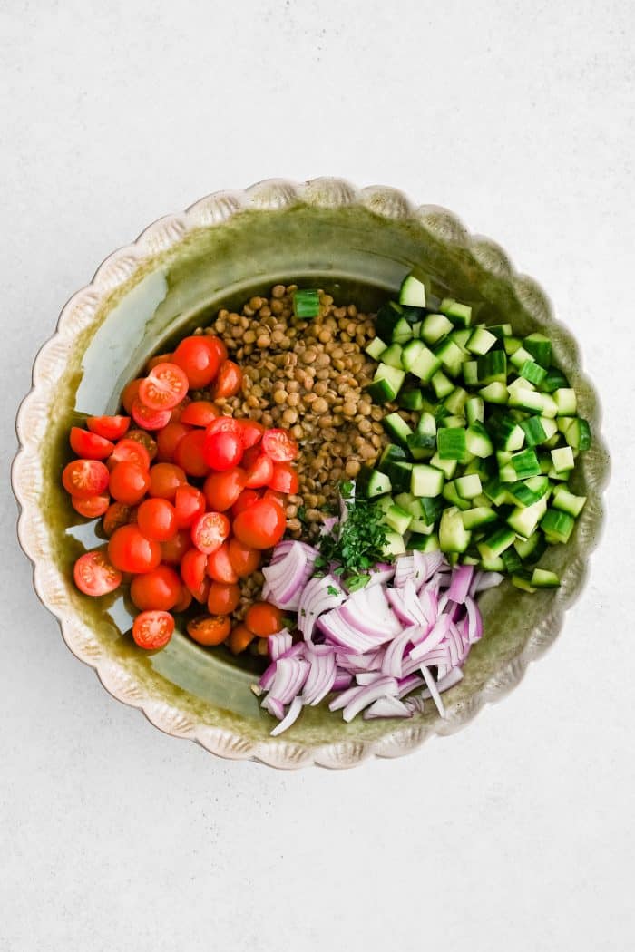 Overhead photo of a scalloped light green serving bowl filled with cooked lentils, halved red cherry tomatoes, diced dark green cucumber, sliced purple-red onion, and chopped parsley on a light white background.