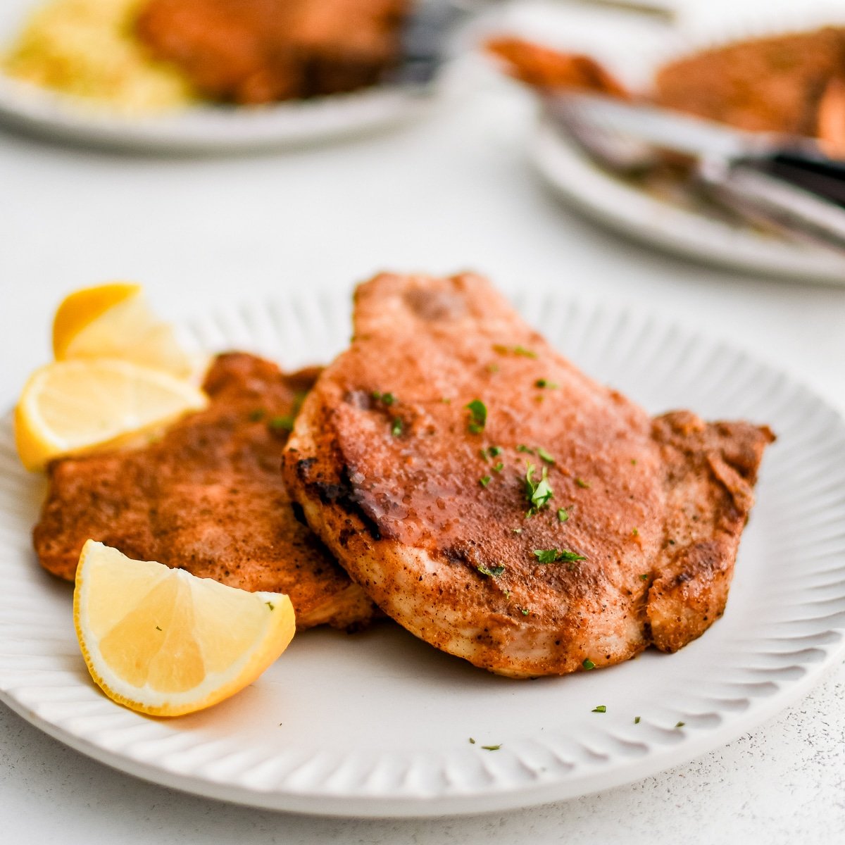 Two perfectly seasoned, golden bone-in pork chops on a white plate with lemon wedges.