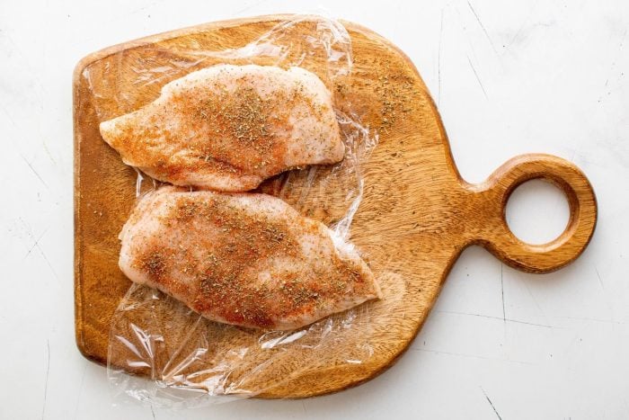 Seasoning two chicken breasts on a cutting board set on top of a piece of plastic wrap.