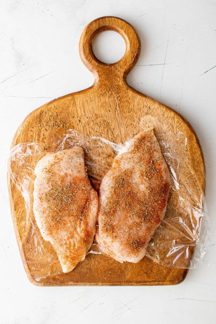 Seasoning two chicken breasts on a cutting board set on top of a piece of plastic wrap.