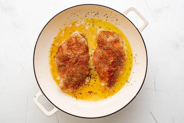 Two well-seasoned chicken breasts pan-frying in a large skillet.