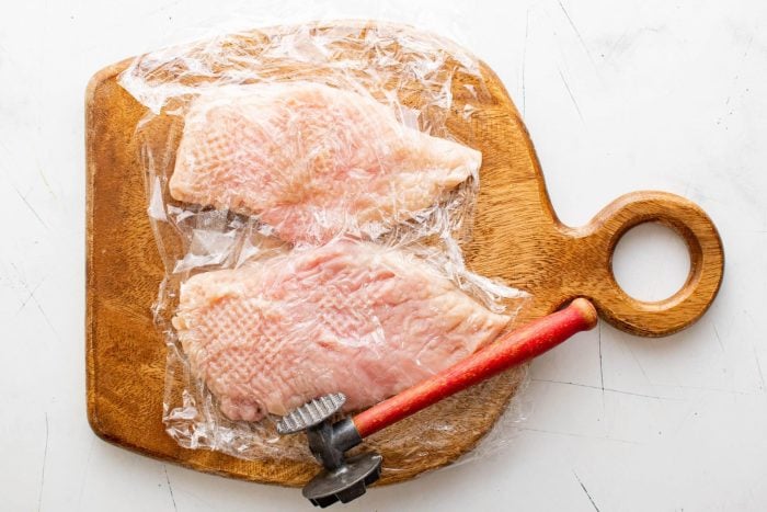Pounding two chicken breasts on a cutting board set between two pieces of plastic wrap.