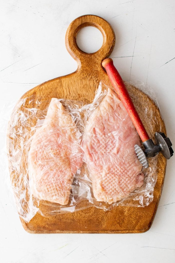 Pounding two chicken breasts on a cutting board set between two pieces of plastic wrap.