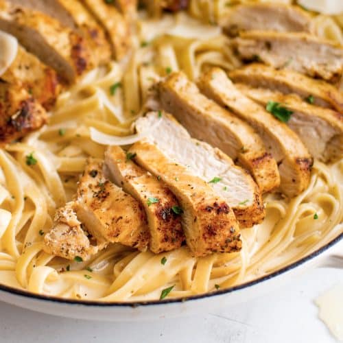 Large skillet with cooked fettuccine pasta coated in creamy Alfredo sauce, topped with two sliced chicken breasts and garnished with freshly grated parmesan cheese.