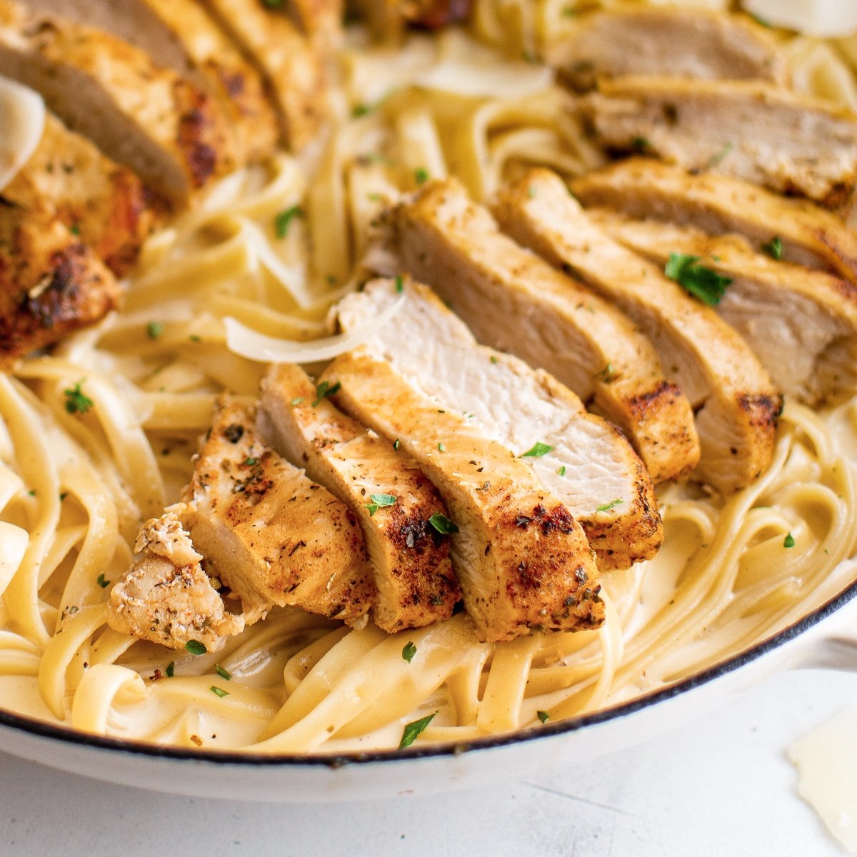 Large skillet with cooked fettuccine pasta coated in creamy Alfredo sauce, topped with two sliced chicken breasts and garnished with freshly grated parmesan cheese.