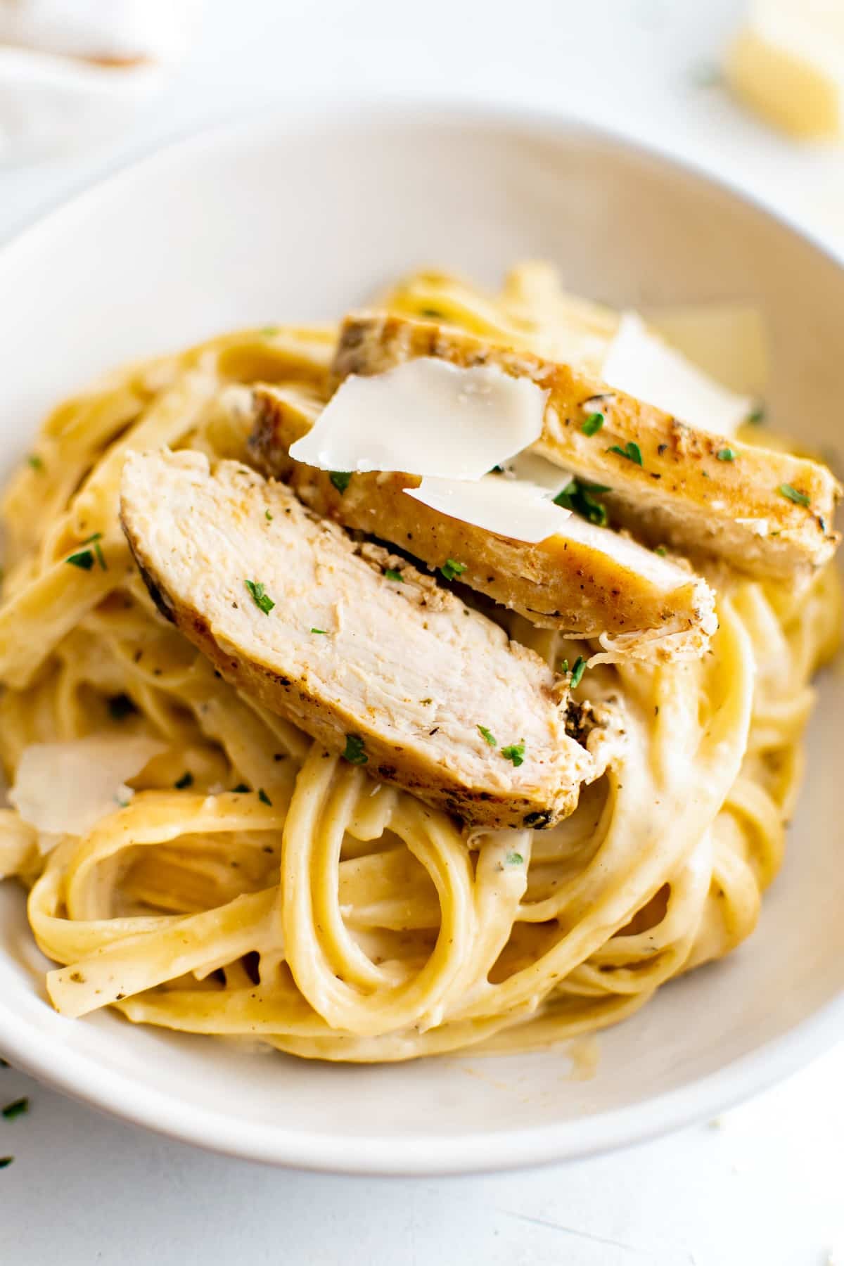 Shallow white serving bowl filled with cooked fettuccine pasta coated in creamy Alfredo sauce and topped with sliced chicken and freshly shredded parmesan cheese.
