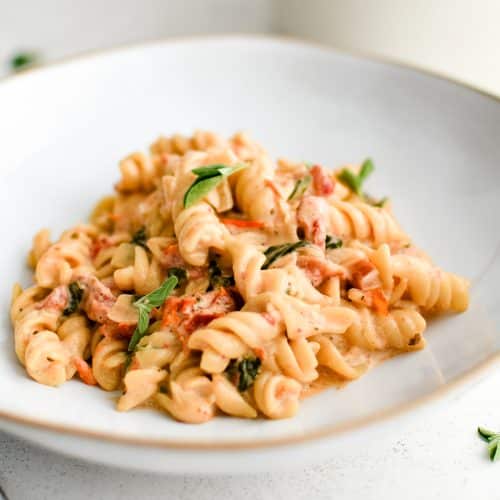 Large shallow plate filled with cooked rotini pasta coated in a homemade creamy sun-dried tomato sauce.