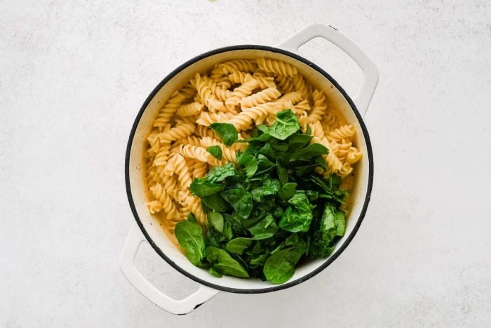 Cooked pasta and baby spinach added to a large pot filled with creamy sun-dried tomato sauce.
