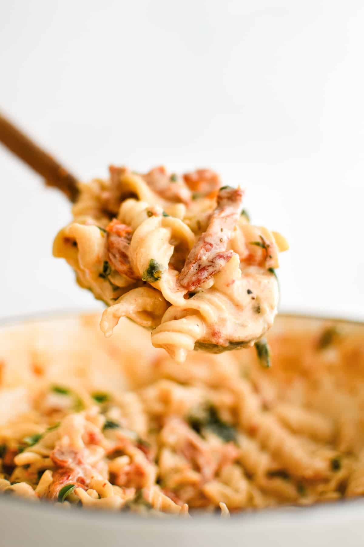 Wooden spoon scooping a large helping of creamy sun-dried tomato pasta from a large pot.