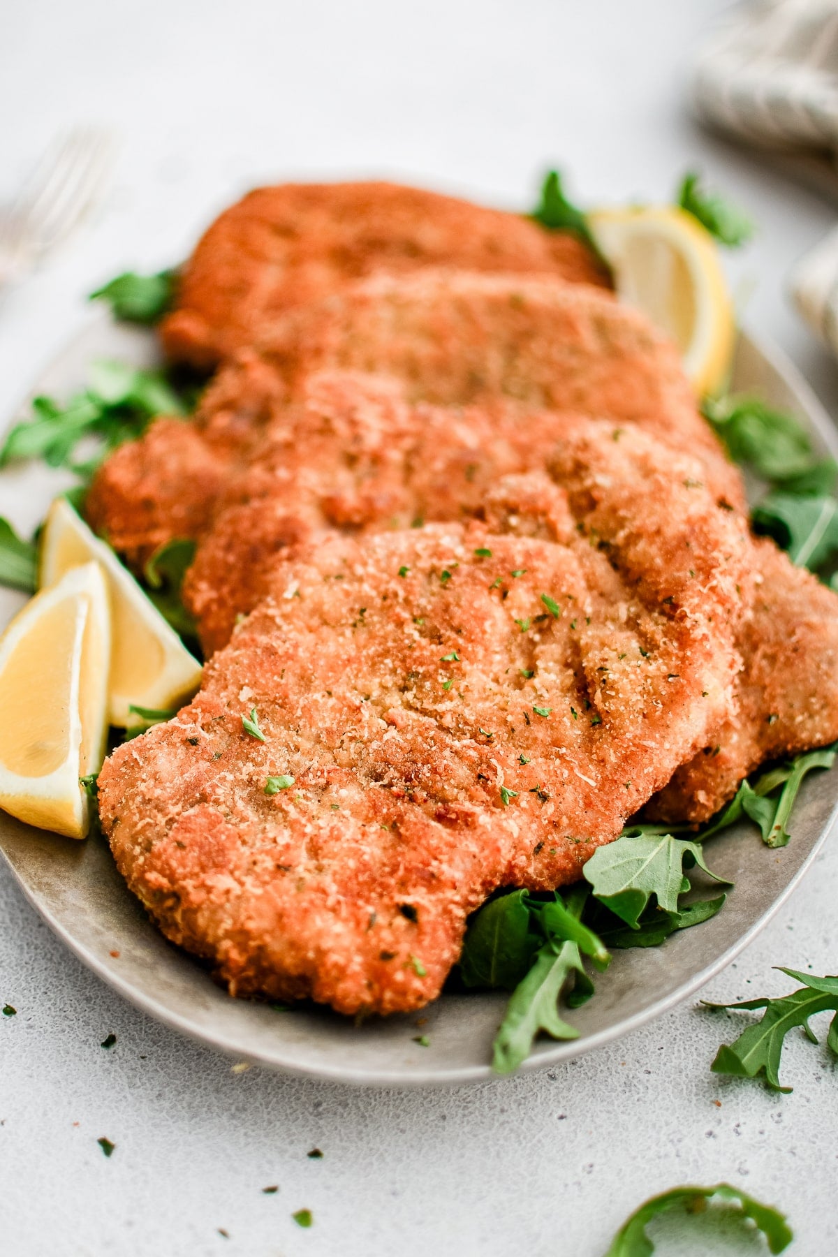 White plate with four perfectly crispy, golden pork Milanese garnished with chopped parsley, lemon, and a light salad of arugula and shaved parmesan.