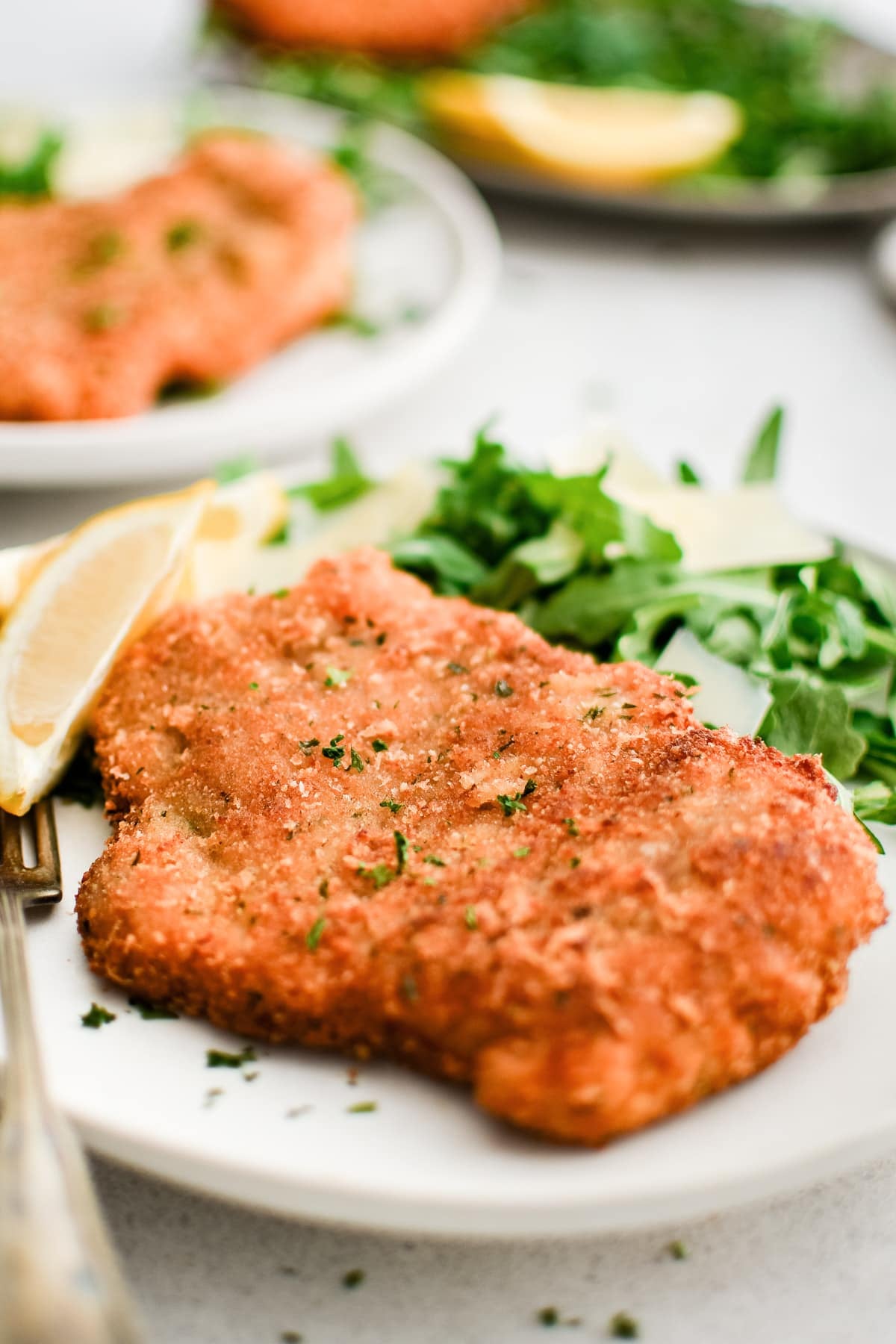 White plate with perfectly crispy, golden pork Milanese garnished with chopped parsley, one wedge of lemon, and a light salad of arugula and shaved parmesan.