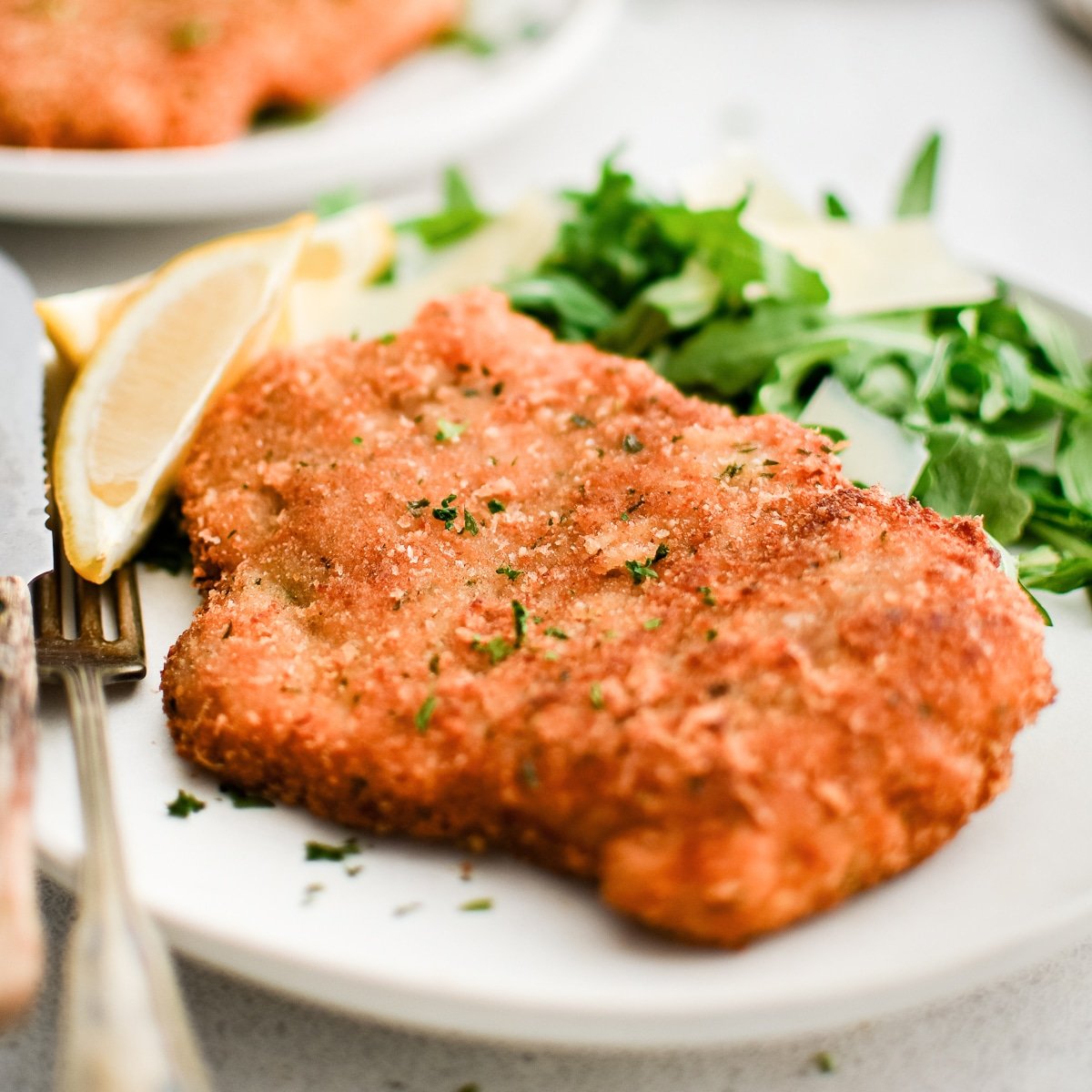 White plate with perfectly crispy, golden pork Milanese garnished with chopped parsley, one wedge of lemon, and a light salad of arugula and shaved parmesan.