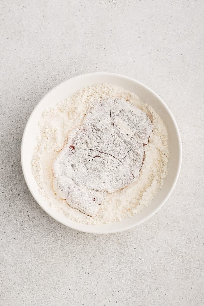 Large flattened pork cutlet in a white bowl filled with flour.