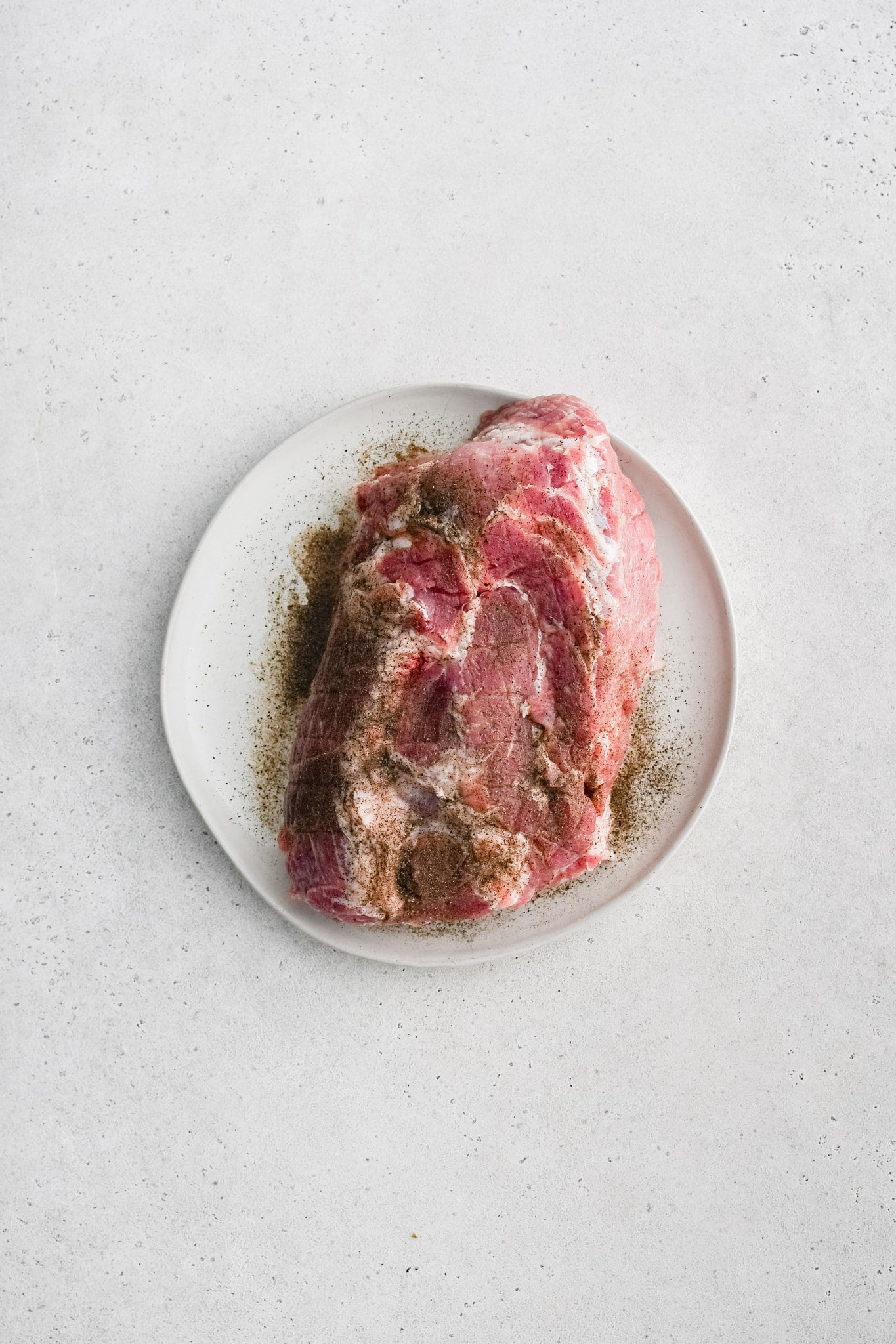 Large boneless pork butt on a white plate seasoned with salt and black pepper.