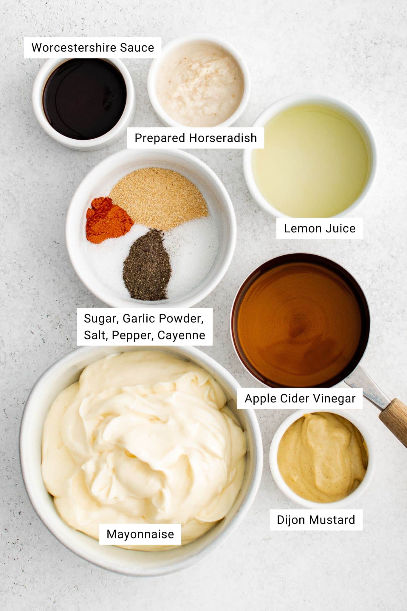 Overhead view of ingredients laid out on a kitchen counter — a bowl of mayonnaise, small bowls of spices, vinegar, mustard, lemon juice, and horseradish.
