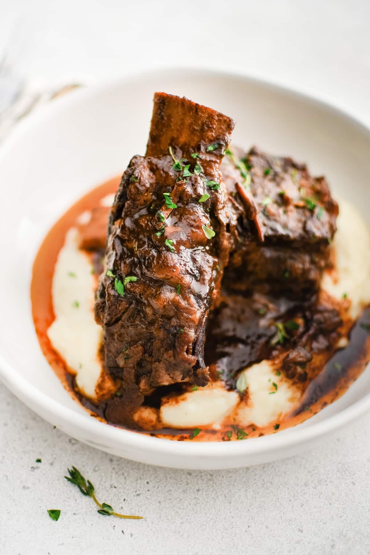 A shallow white bowl filled with creamy mashed potatoes and topped with two slow cooked beef short ribs smothered in red wine sauce and garnished with fresh thyme.