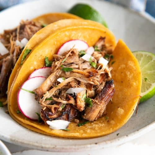 Two pork carnitas tacos served on warm corn tortillas, topped with fresh cilantro, diced onions, sliced radish, and a squeeze of lime, presented on a shallow serving plate.