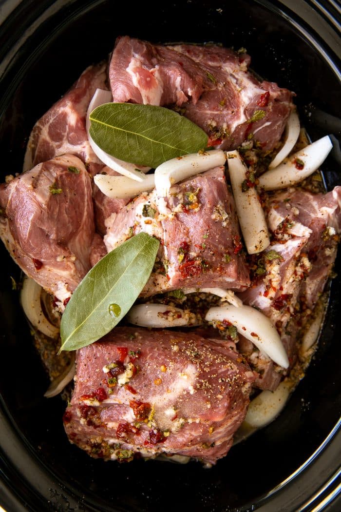 Large chunks of pork butt in a slow cooker covered by sliced onion, bay leaves, and a flavorful seasoning broth made with Mexican oregano, ground cumin, minced garlic, chili powder, orange juice, lime juice, chipotles in adobo, and chicken broth (or light Mexican beer).