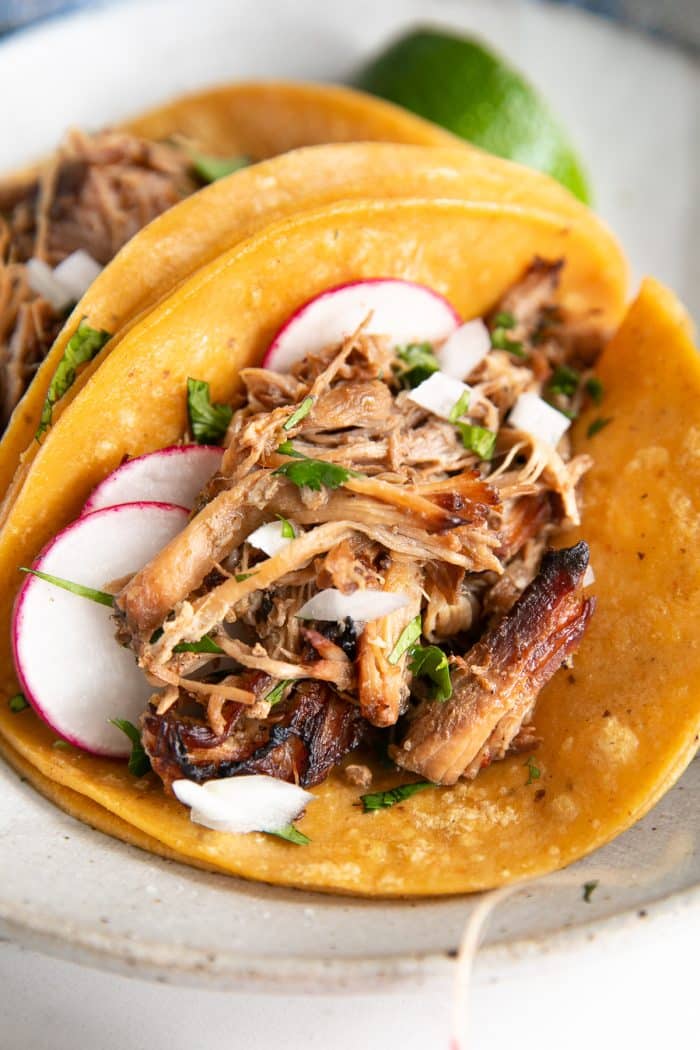 Two pork carnitas tacos served on warm corn tortillas, topped with fresh cilantro, diced onions, sliced radish, and a squeeze of lime, presented on a shallow serving plate.