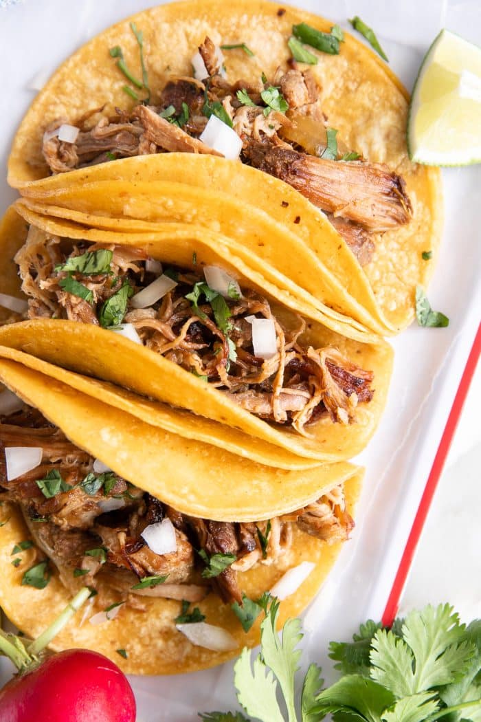 Three pork carnitas tacos on soft corn tortillas, filled with tender, crispy shredded pork, garnished with fresh cilantro and diced onions, served with lime wedges on the side.