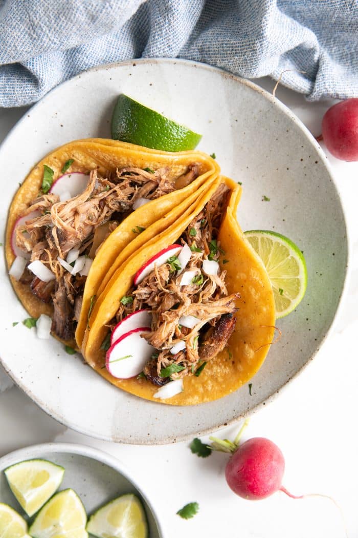 Two pork carnitas tacos served on warm corn tortillas, topped with fresh cilantro, diced onions, sliced radish, and a squeeze of lime, presented on a shallow serving plate.