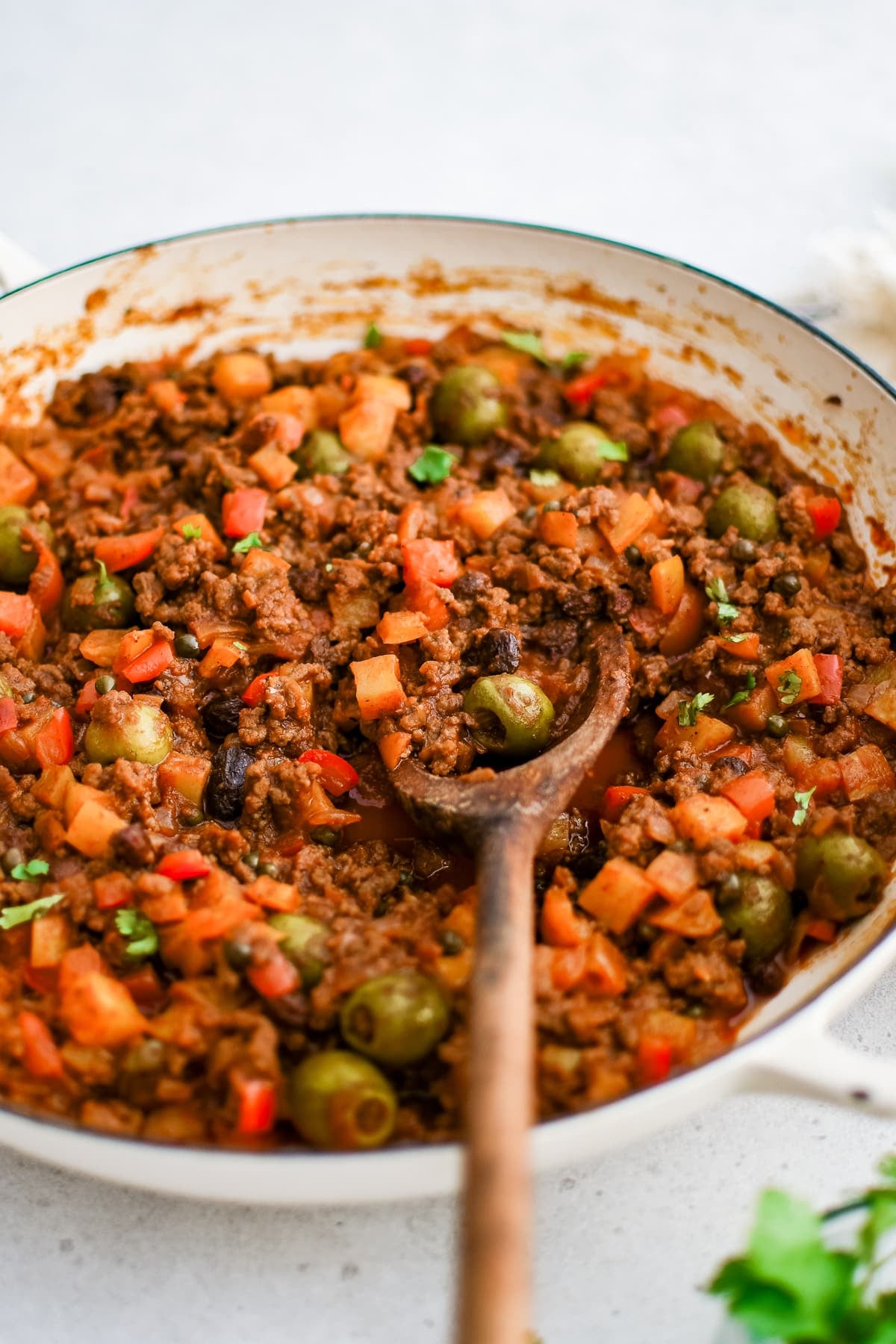 A wooden serving spoon rests inside a simmering pot of Cuban picadillo with ground beef, olives, raisins, and potatoes in a rich tomato sauce.