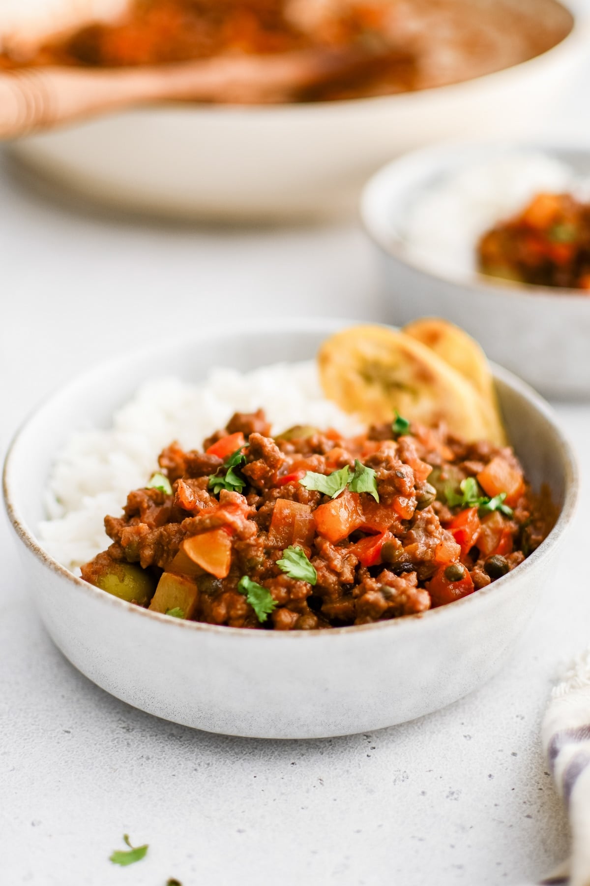 A bowl of Cuban picadillo filled with ground beef, red peppers, olives, and potatoes, served with white rice and fried plantains.
