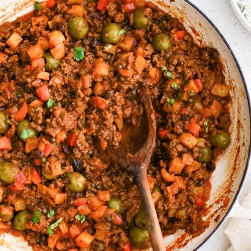 A wooden serving spoon rests inside a simmering pot of Cuban picadillo with ground beef, olives, raisins, and potatoes in a rich tomato sauce.