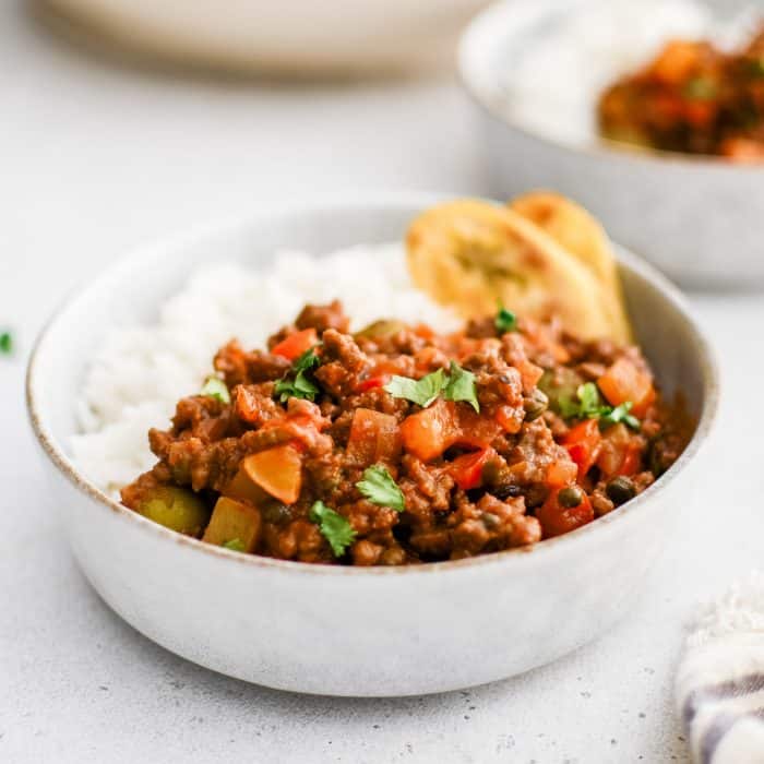 A bowl of Cuban picadillo filled with ground beef, red peppers, olives, and potatoes, served with white rice and fried plantains.