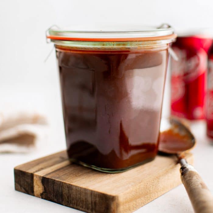 Homemade Dr. Pepper BBQ sauce in a glass storage jar with a spoon set to its left and a Dr. Pepper can in the background.