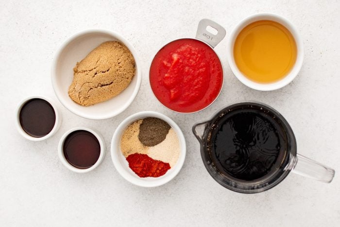 Ingredients for Dr. Pepper BBQ sauce including Dr. Pepper, tomato sauce, brown sugar, vinegar, spices, and Worcestershire sauce measured out into individual bowls ready to be used.