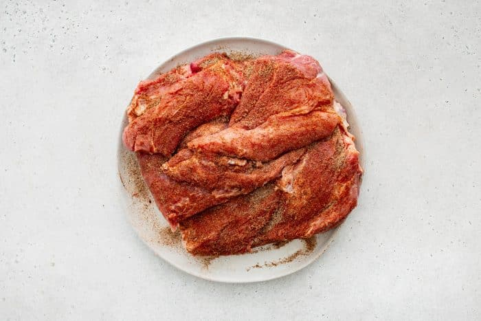A raw pork shoulder on a white plate is generously rubbed with a blend of salt, pepper, garlic powder, onion powder, and smoked paprika.