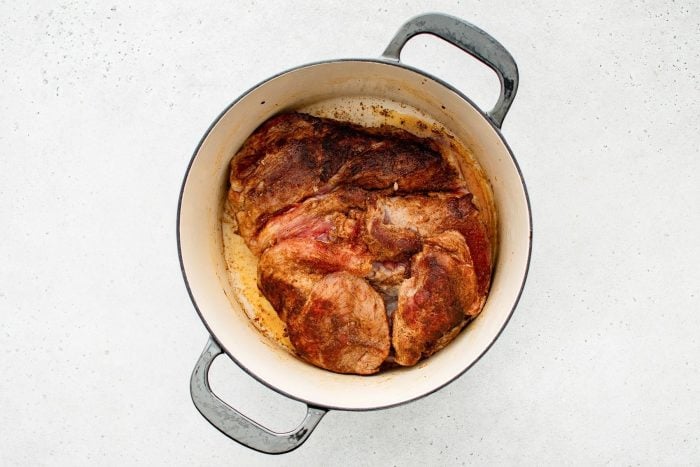 A pork shoulder sizzling in a Dutch oven, forming a golden-brown crust as it sears in olive oil.