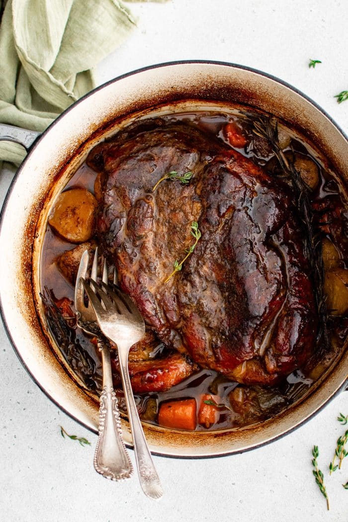 Two forks rest in a large Dutch oven filled with a perfectly cooked fork-tender pork roast surrounded by tender vegetables.