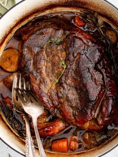 Two forks rest in a large Dutch oven filled with a perfectly cooked fork-tender pork roast surrounded by tender vegetables.