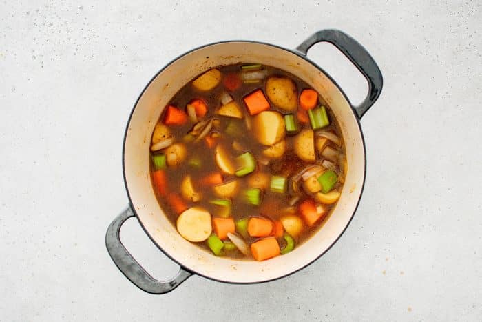 A seared pork roast nestled in broth, surrounded by chopped carrots, potatoes, celery, and fresh sprigs of rosemary and thyme.