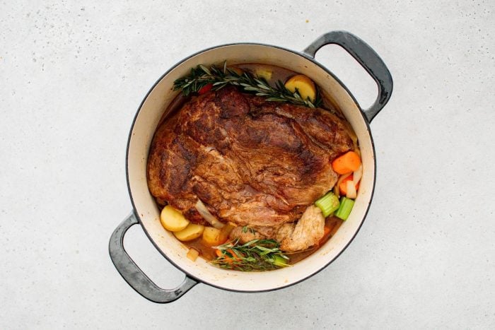A seared pork roast nestled in broth, surrounded by chopped carrots, potatoes, celery, and fresh sprigs of rosemary and thyme.