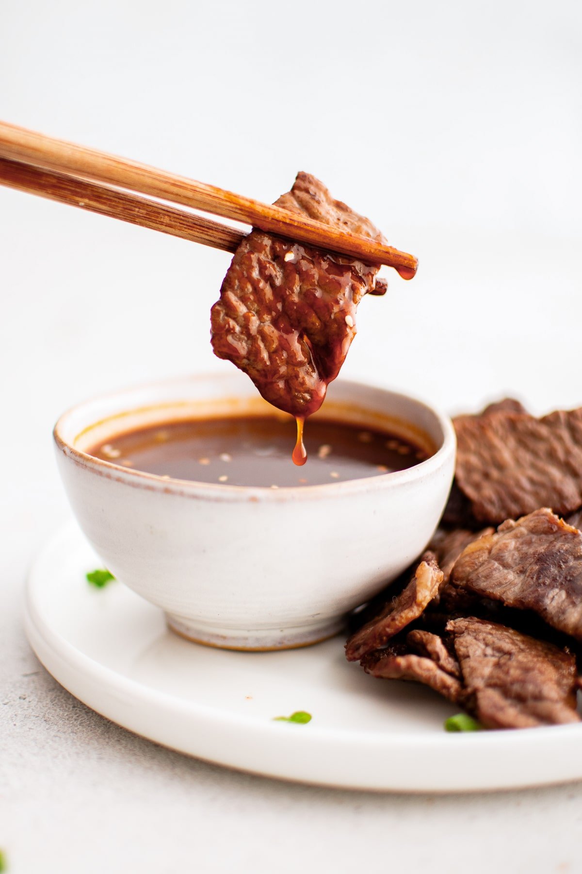 A slice of thinly grilled beef held by chopsticks being dipped into a small bowl of yakiniku sauce.