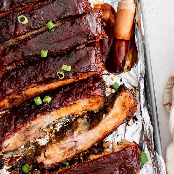 Oven baked sliced spare ribs glazed with BBQ sauce and topped with green onions resting on a foil-lined pan with a red-tipped basting brush beside them.