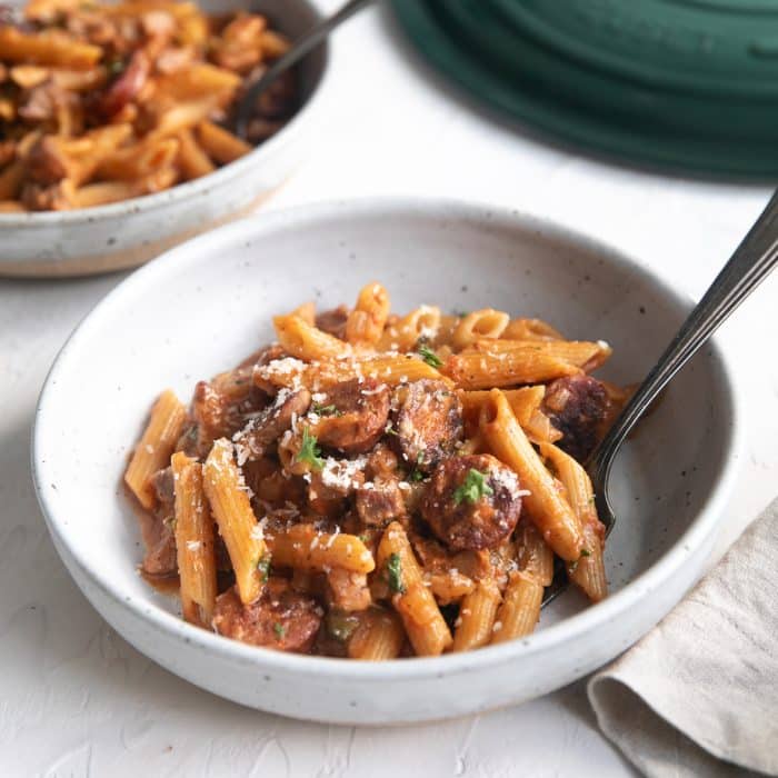 A shallow white plate holds a hearty serving of Pastalaya. The pasta is coated in a vibrant reddish-orange sauce, flecked with herbs and bits of meat. The dish is garnished with finely chopped parsley and a light dusting of grated Parmesan, adding contrast and freshness.