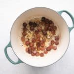 Rounds of smoked andouille sausage are sizzling in a Dutch oven, some pieces beginning to brown and crisp at the edges. A small amount of rendered fat glistens at the bottom of the pot, creating a flavorful base.