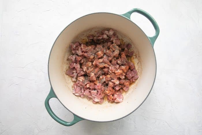 Small cubes of pork shoulder seasoned with salt, black pepper, and Cajun seasoning added to a large green-handled Dutch Oven.