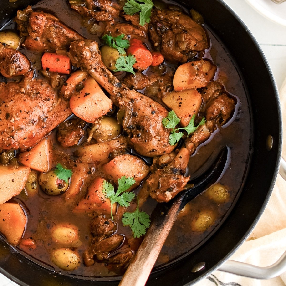 A large pot of Puerto Rican Pollo Guisado simmering with bone-in chicken, potatoes, carrots, and green olives in a rich tomato-based sauce.