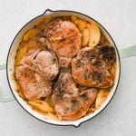 Pork chops simmering in a skillet with apples, onions, and sauce