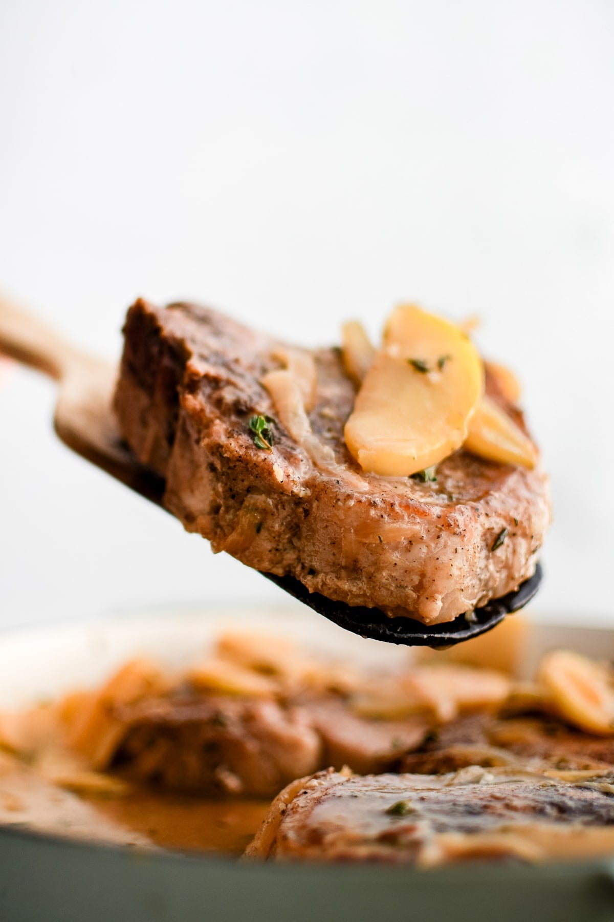 A wooden spatula holding a seared pork chop above a skillet filled with apples and onions.