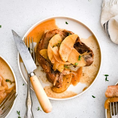 Pork chop served on a white plate with apples, onions, and sauce drizzled on top.