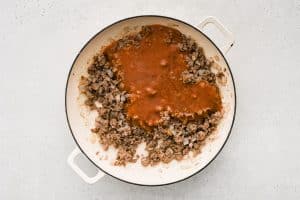 Homemade sloppy joe sauce poured into a large skillet filled with browned ground beef and sautéed onions.